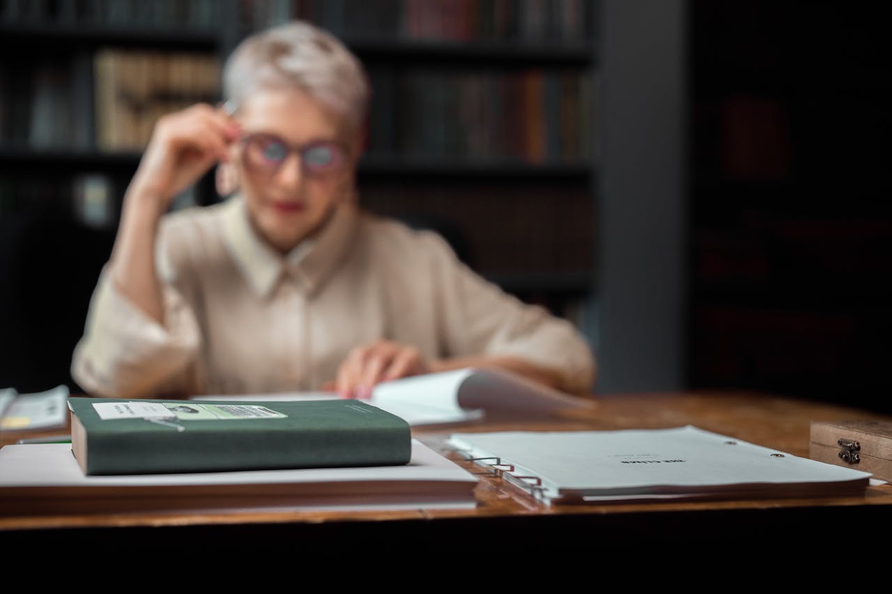 Senior woman in library setting, focused on reading with a book and documents in foreground.